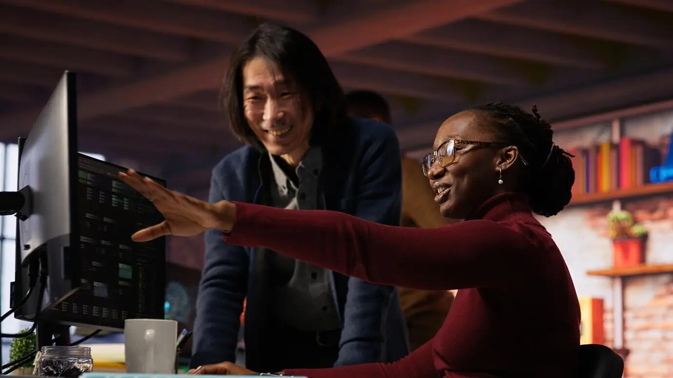 Довольный руководитель команды шутит с сотрудником, занимающимся тестированием кода, в непринужденной рабочей обстановке.